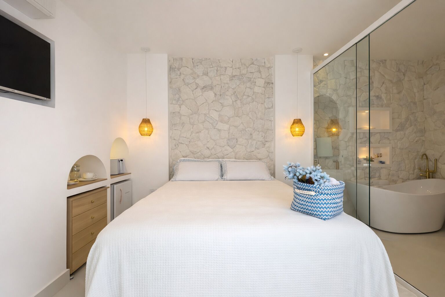 A modern bedroom with a large bed, a stone accent wall, a glass shower enclosure, and a bedside table with a lamp.
