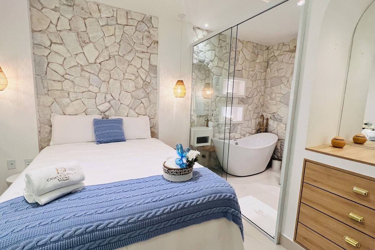 Un dormitorio moderno con una pared de piedra, una cama grande con manta azul, una mesita de noche y un baño con bañera y espejo.