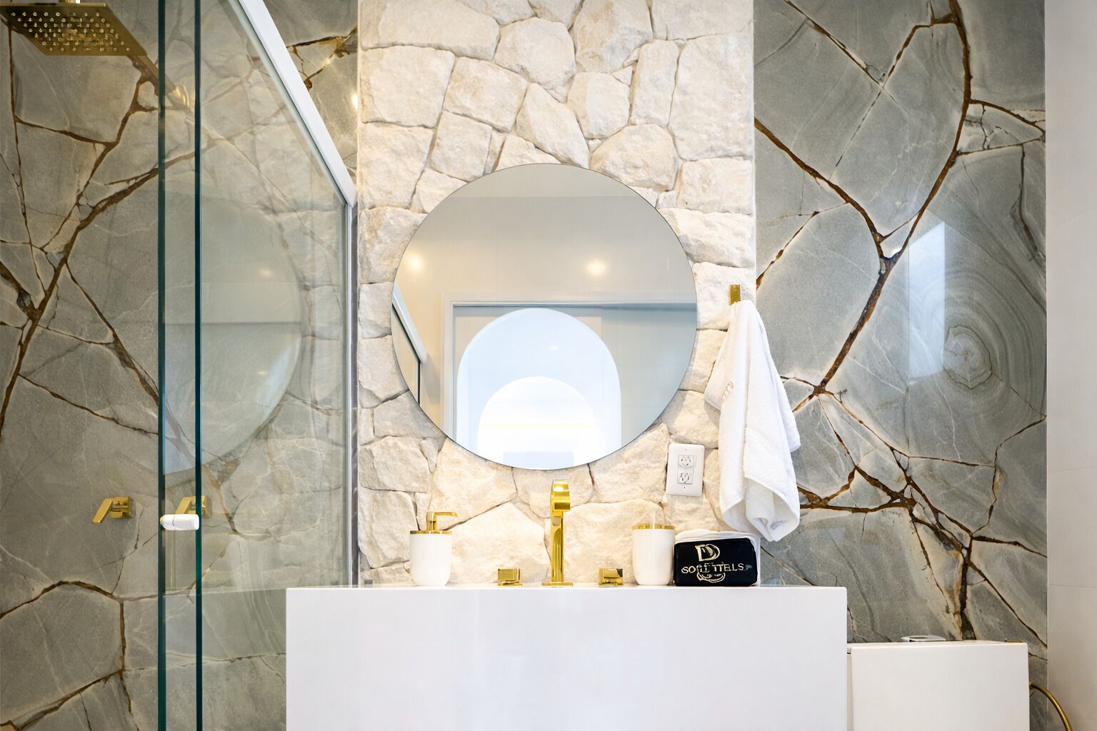 A bathroom with a stone wall, a round mirror, a white sink, and a glass shower enclosure.