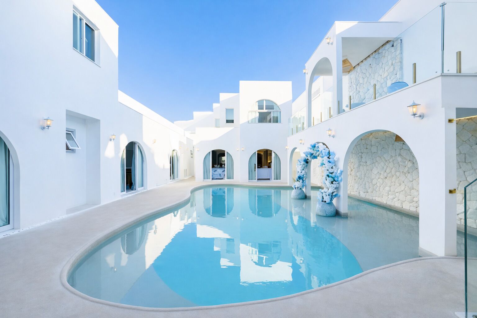 Zona de piscina con edificios blancos, arcos y un cielo azul despejado.