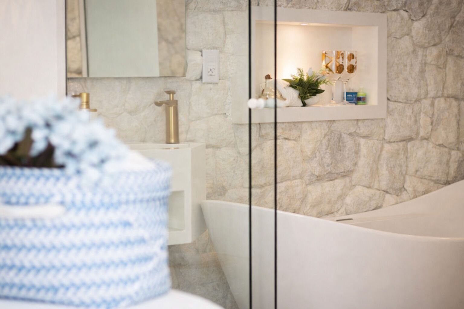 A modern bathroom with a stone wall, a white sink, a gold faucet, and a white bathtub.