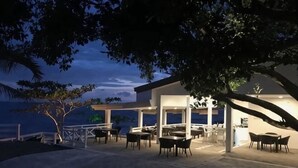Ένας εξωτερικός χώρος με τραπέζια και καρέκλες, μια σκεπαστή κατασκευή και θέα στη θάλασσα.