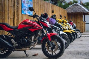 A row of motorcycles parked outside a building with a wooden fence and a thatched-roof structure in the background. - Cliffside Resort (Panglao Municipality)