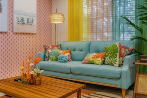 Un salon avec un canapé bleu clair, des coussins aux couleurs vives sur le thème tropical, une table basse en bois ornée de vases et une lampe fixée au mur.