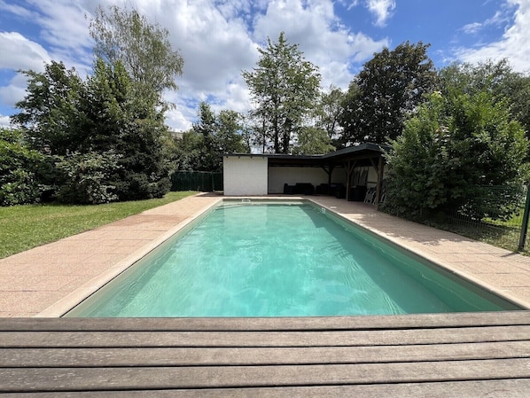 A rectangular swimming pool with a wooden deck in the foreground, surrounded by a grassy area and trees.