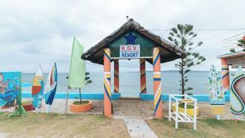 Entrada de un complejo turístico frente al mar con un arco de colores, un letrero que dice R.G.V. Resort y tablas de surf a la vista.