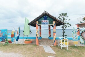 L'entrée d'un complexe hôtelier situé en bord de mer, avec une arche colorée, une enseigne indiquant « R.G.V. Resort », et des planches de surf exposées.