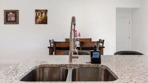 Eine Küche mit Granit-Arbeitsplatten, Edelstahlspülen und einem modernen Wasserhahn. An der Wand hängen zwei gerahmte Bilder, und es gibt einen Essbereich mit Holztischen und einer Tischplatte.