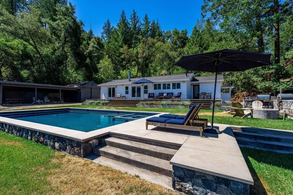 A swimming pool with steps leading to a deck area, a covered patio, and a house in the background.