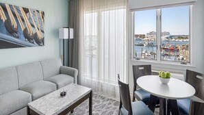 Une chambre d’hôtel moderne avec un canapé gris, une table basse en marbre, une table à manger avec des chaises, une grande fenêtre donnant sur un port de plaisance et des bateaux, et un tableau accroché au mur.