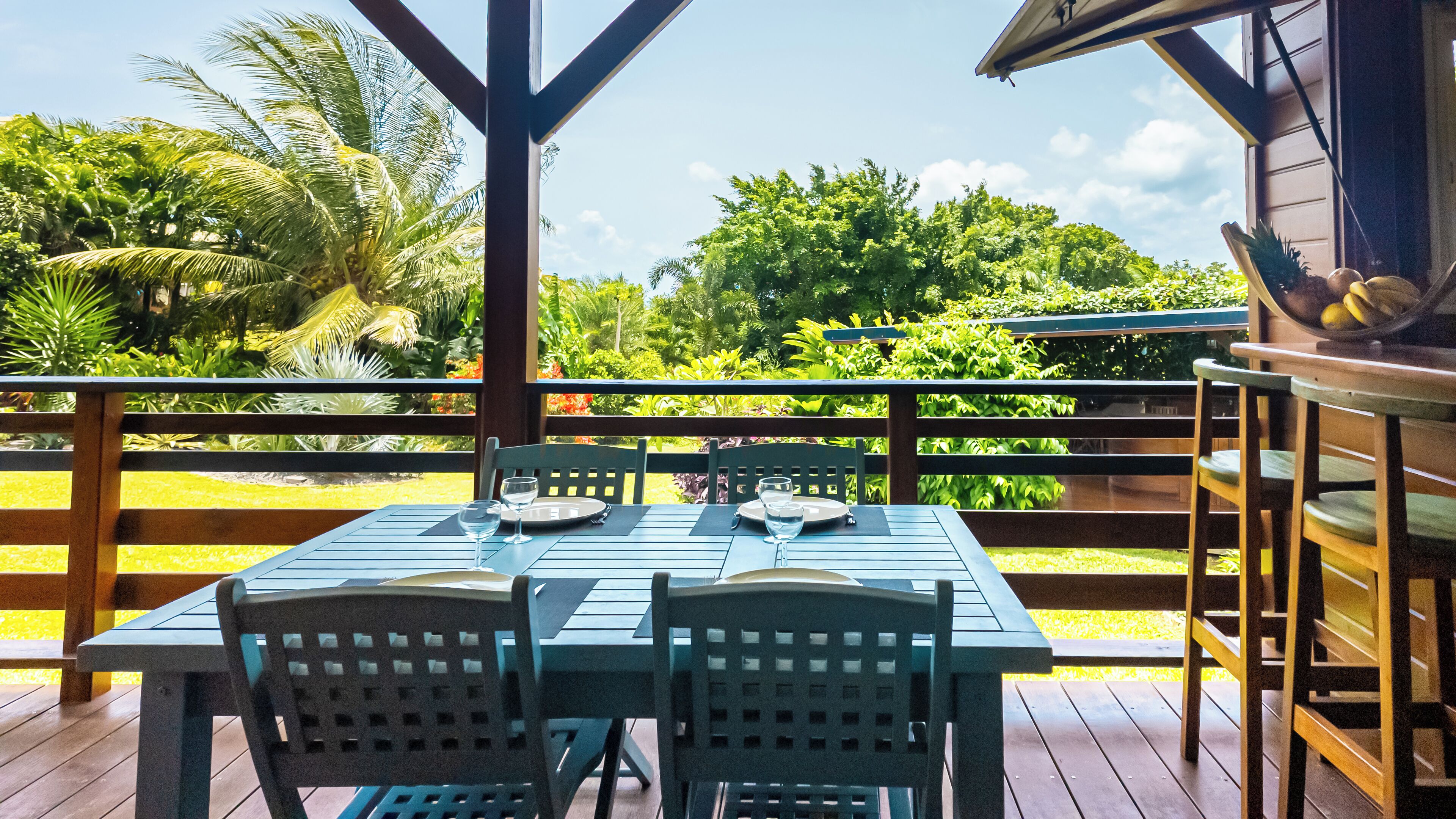 A wooden deck with a table set for two, overlooking a lush garden with various plants and trees.