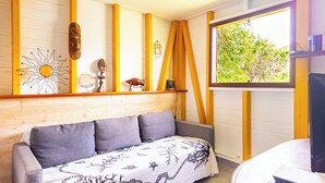 A cozy room with a wooden wall, a sofa with a patterned cover, a window with a view of greenery, and a shelf with various items. - Wooden bungalow Ti Briz - Au jardin d'Éole (Sainte-Rose)