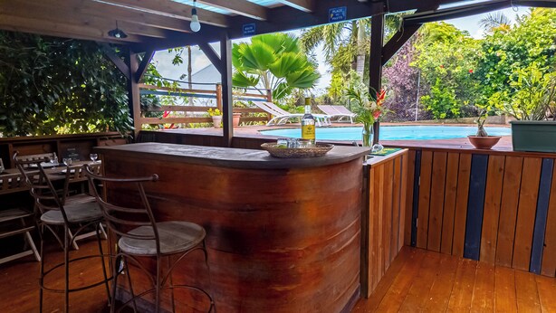 An outdoor bar area with wooden furniture, a pool in the background, and various potted plants. - Wooden bungalow Ti Briz - Au jardin d'Éole (Sainte-Rose)