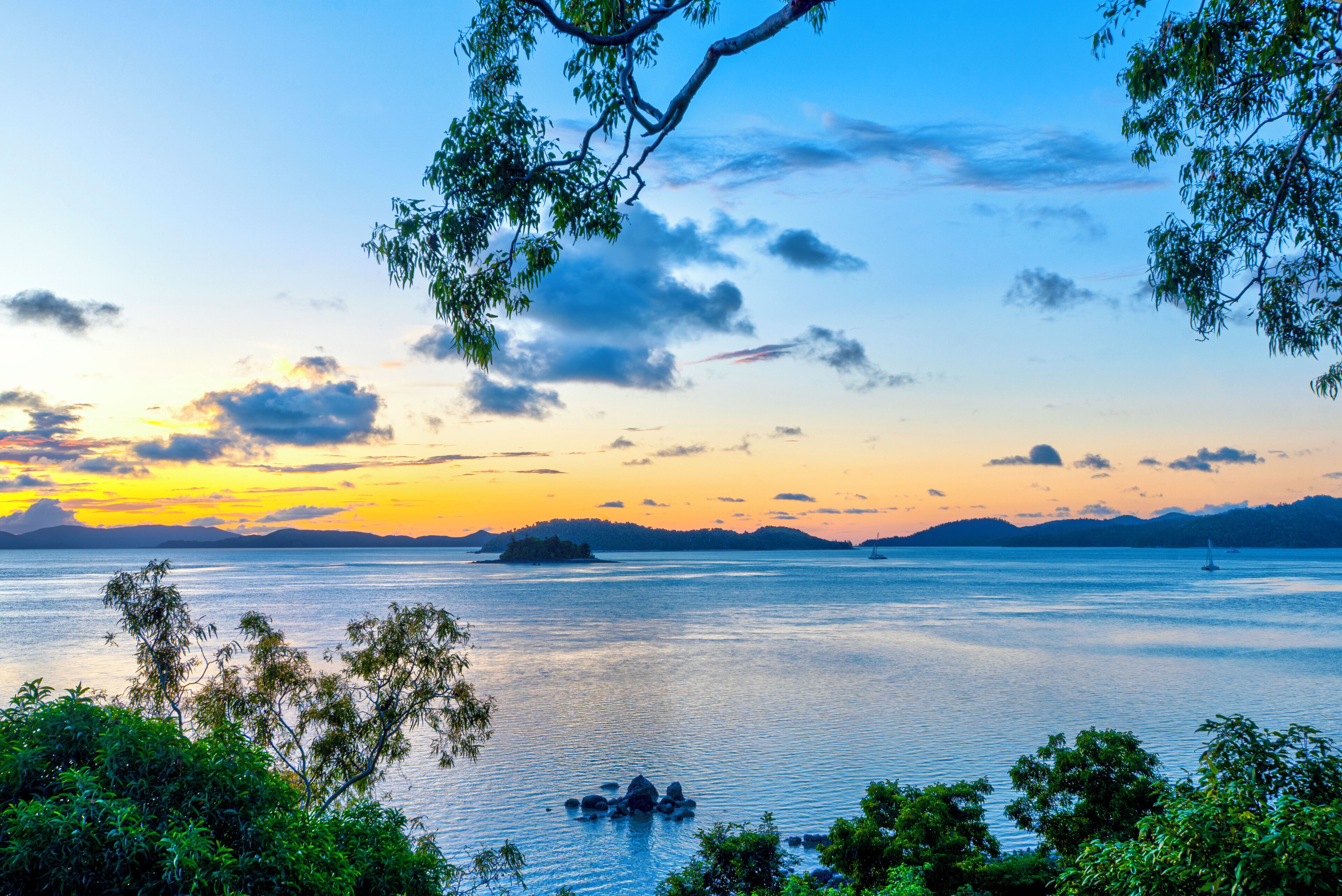 A serene lake at sunset with a distant island and a sailboat.
