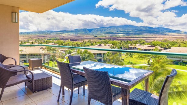 A balcony with wicker furniture and a glass table, overlooking a scenic landscape with mountains and greenery.
