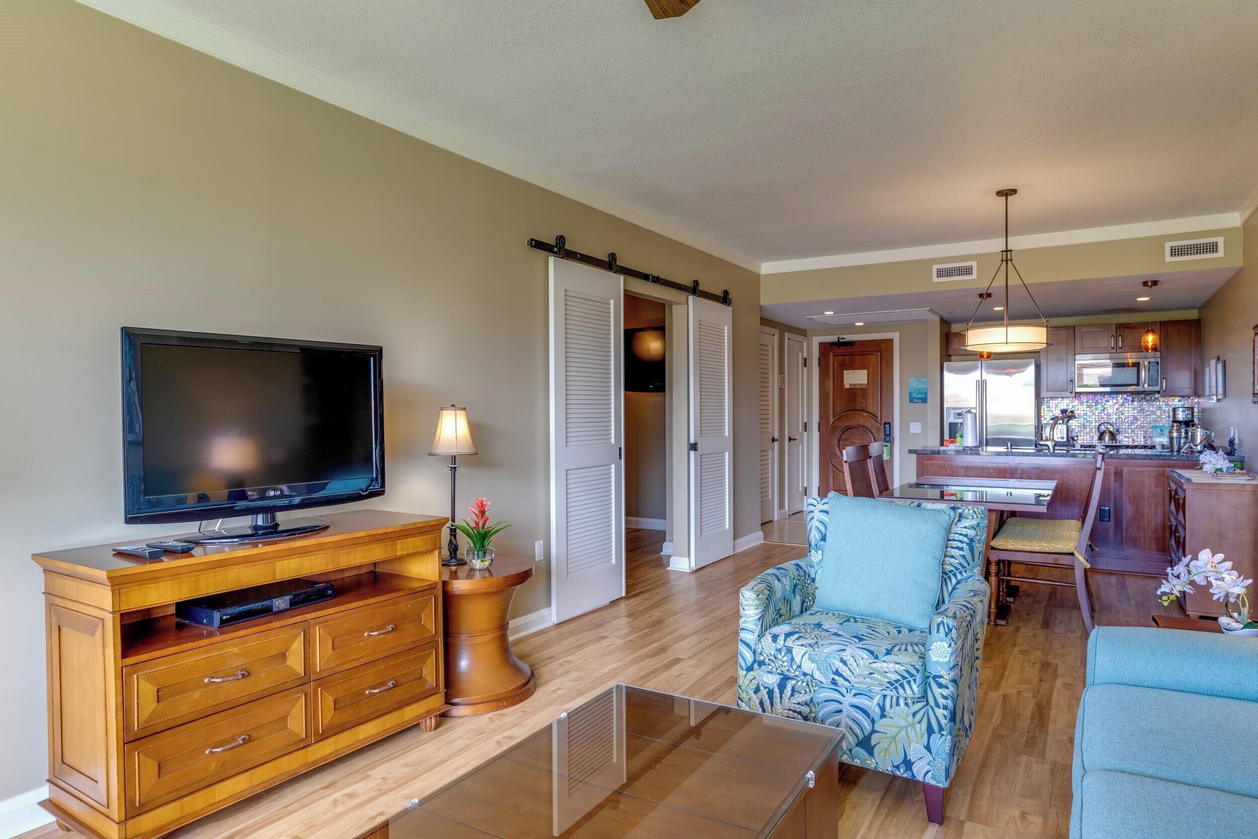 A living room with a flat-screen TV, wooden entertainment unit, and a patterned armchair. A kitchen with wooden cabinets and a dining area is visible in the background.