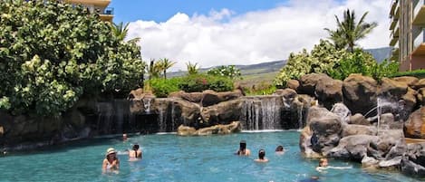 A swimming pool area with multiple waterfalls and large rocks, surrounded by lush greenery and a clear sky.