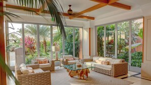 A spacious living room with wicker furniture, large windows, and a view of lush greenery.
