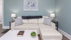 Una sala de estar moderna con una pared azul claro, un sofá seccional blanco, dos mesas auxiliares con lámparas, una mesa de centro con un libro y una planta, y un cuadro de flores en la pared.
