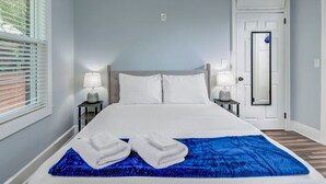 Una cama bien hecha con sábanas blancas y una manta azul, dos lámparas de noche, una puerta con espejo y una ventana con persianas.