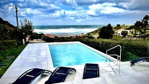A swimming pool with two sun loungers, a view of the ocean, and a hilly landscape in the background. - Balcobo Villas&Cabañas (Arteixo)