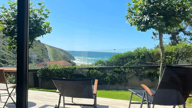 A room with a large window overlooking a coastal view, featuring two wicker chairs and a bed with patterned bedding. - Balcobo Villas&Cabañas (Arteixo)