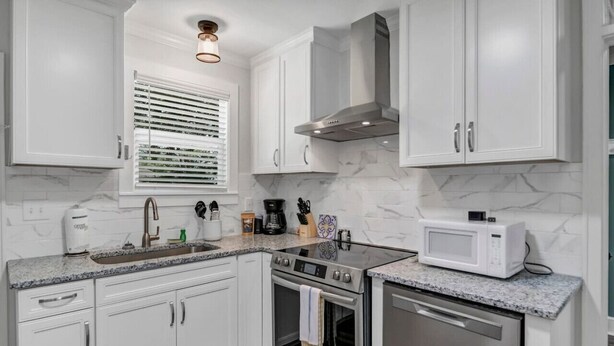 A modern kitchen with white cabinets, a stainless steel range, a microwave, and a window with blinds. - Capes Cottage | 1BR Near Rosewood (Columbia)
