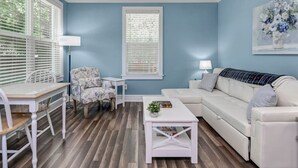 A living room with a blue wall, white furniture, a white sofa, a white coffee table, a white chair, and a window with blinds. - Capes Cottage | 1BR Near Rosewood (Columbia)