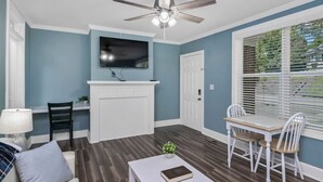 A living room with a fireplace, a ceiling fan, a TV mounted above it, and a dining area with a table and chairs. - Capes Cottage | 1BR Near Rosewood (Columbia)
