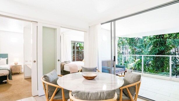 A modern, well-lit room with a round table and chairs, a large window with a view of greenery, and a sliding glass door leading to another room.