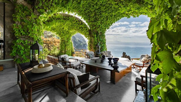 A cozy outdoor seating area with a table and chairs, surrounded by lush green vines and a view of the sea.