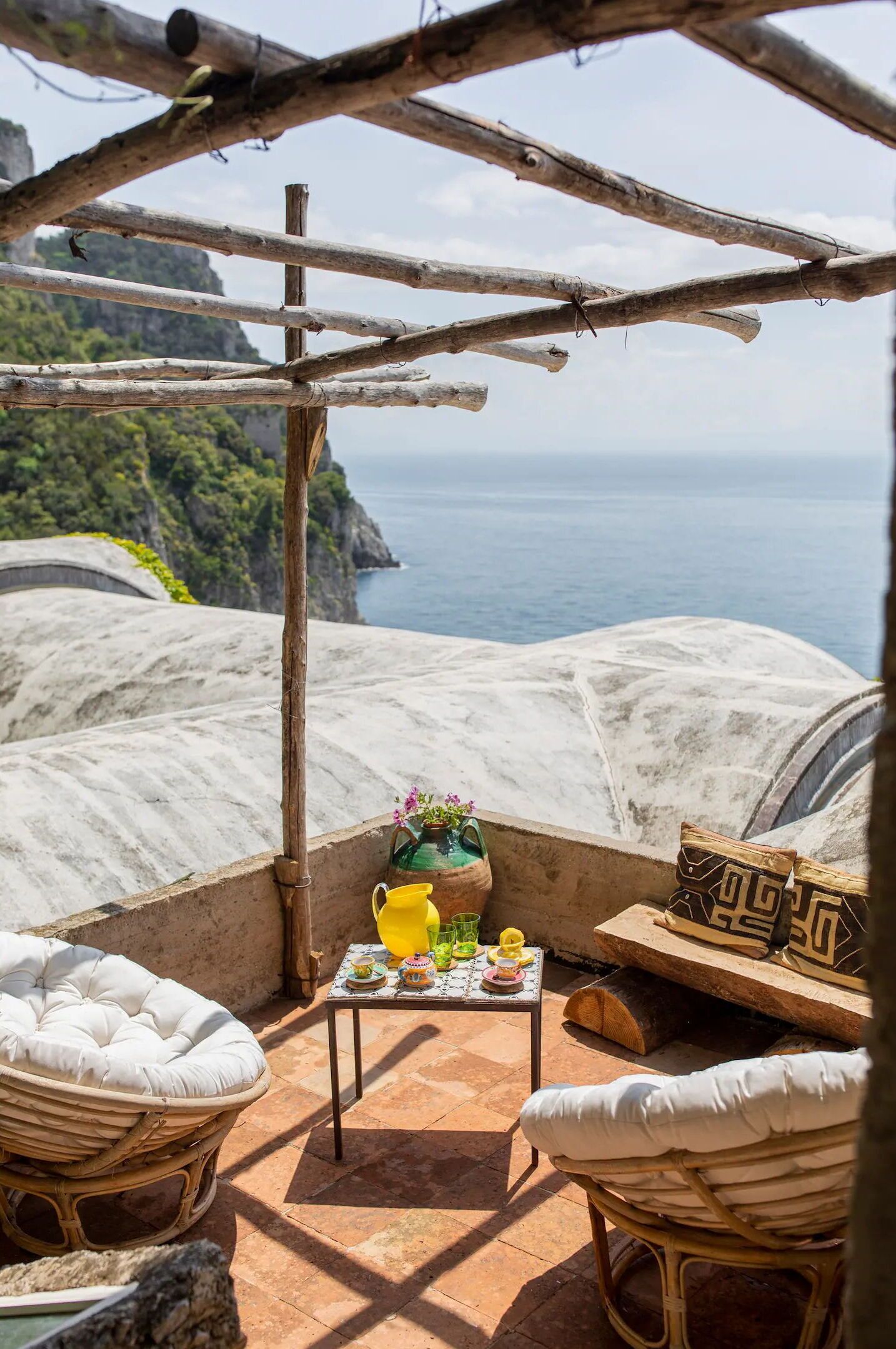 A terrace with wicker furniture, a small table with a vase and drinks, and a view of the sea and cliffs.