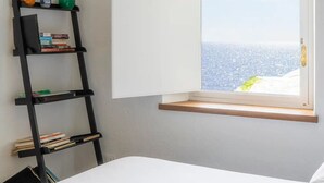 A bedroom with a bed, a window with an open view of the sea, a black ladder, and a shelf with books and decorative items.