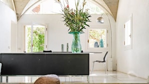 A spacious room with a high arched ceiling, a wooden dining table with a woven basket, a vase with flowers, and a view of greenery outside.