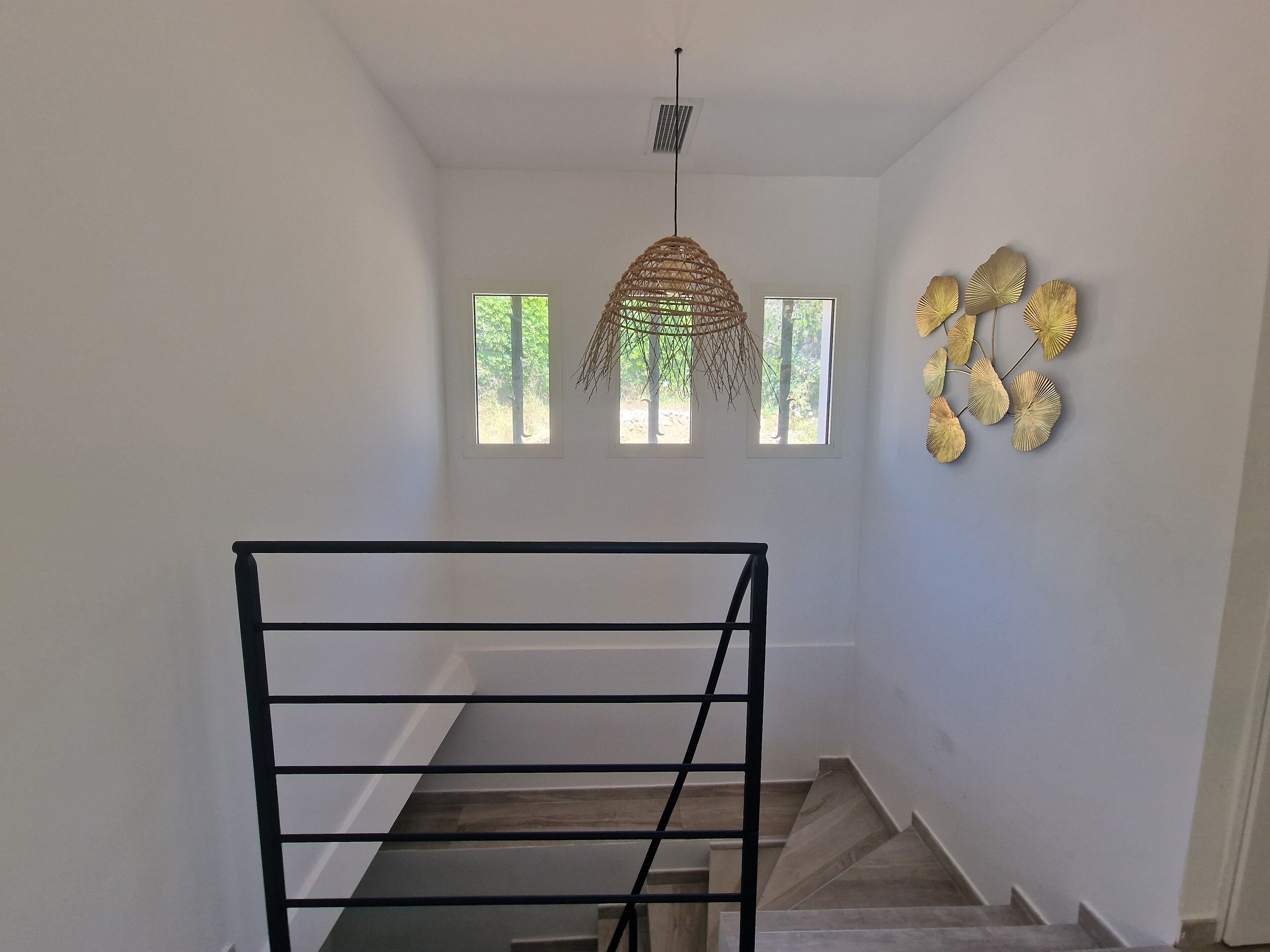 A staircase with a black metal railing leading up to a landing. There are three windows on the landing wall, and a decorative wall hanging with leaf-like patterns to the right.