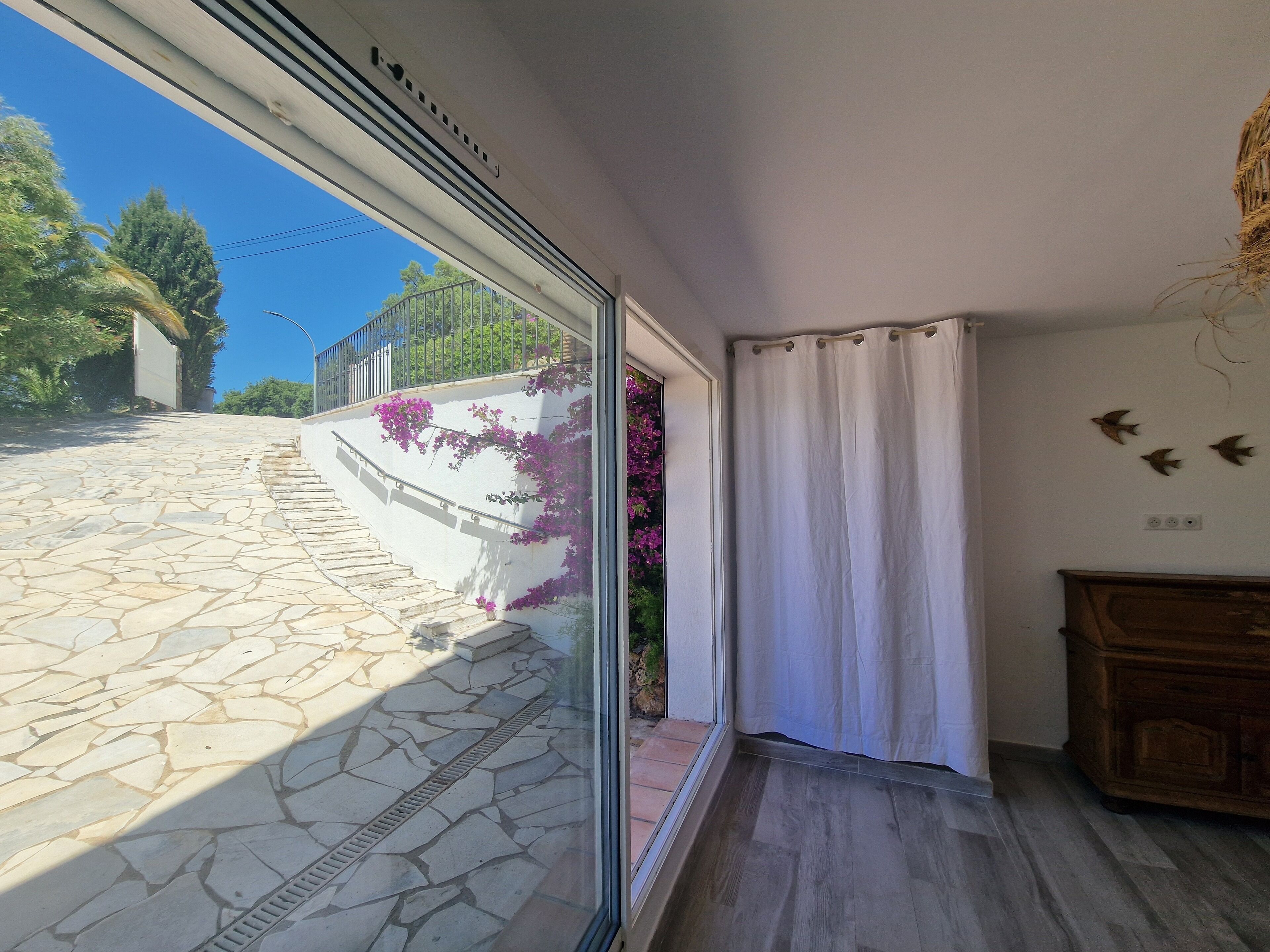 Ein Zimmer mit einer Steintreppe, die nach draußen führt, einem Fenster mit Blick auf eine weiße Wand und violette Blumen sowie einem Holzschrank.