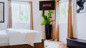 Un dormitorio con cama, planta en maceta, televisor montado en la pared y ventana con cortinas.