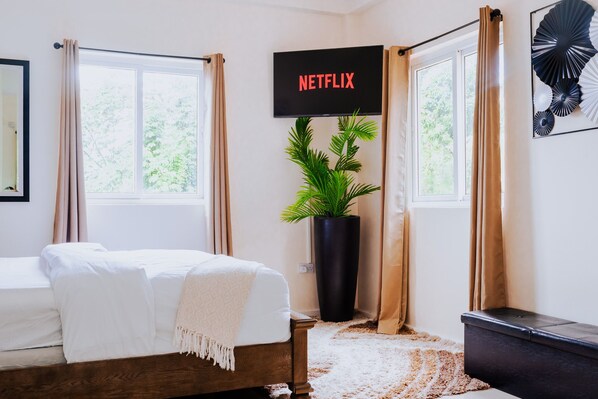 Un dormitorio con cama, planta en maceta, televisor montado en la pared y ventana con cortinas.