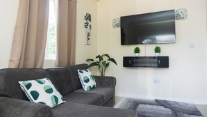 Un salón con un sofá gris, dos cojines blancos y verdes estampados, un televisor de pantalla plana montado en la pared y dos pequeñas macetas con plantas.