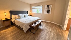A bedroom with a bed, a bench, a nightstand, a lamp, a window, and a painting on the wall.