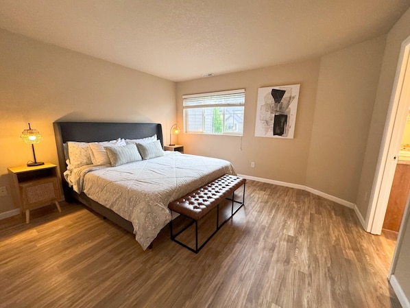 A bedroom with a bed, a bench, a nightstand, a lamp, a window, and a painting on the wall.