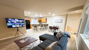 A modern living room with a sectional sofa, a flat-screen TV on a tripod, a dining area with a bar, and a kitchen in the background.