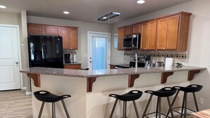 A kitchen with wooden cabinets, a black refrigerator, a microwave, and a granite countertop. There are four black bar stools.