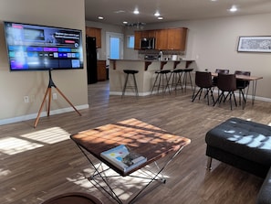 A modern living room with a TV on a stand, a coffee table with magazines, a dining area with a table and chairs, and a kitchen in the background.