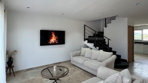 Un salon moderne avec un canapé beige, une table basse en verre, un téléviseur à écran plat et un escalier menant à un niveau supérieur.