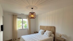 Une chambre spacieuse avec un grand lit, une tête de lit en bois, un luminaire suspendu, une table de chevet ornée d’un vase et une chaise.