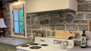 Una cocina con estufa blanca, gabinetes verdes, pared de piedra y ventana con vista a árboles.