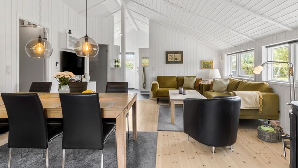 A spacious living area with a wooden dining table, black chairs, a sofa, and a coffee table. There are pendant lights above the table and a lamp on the side.