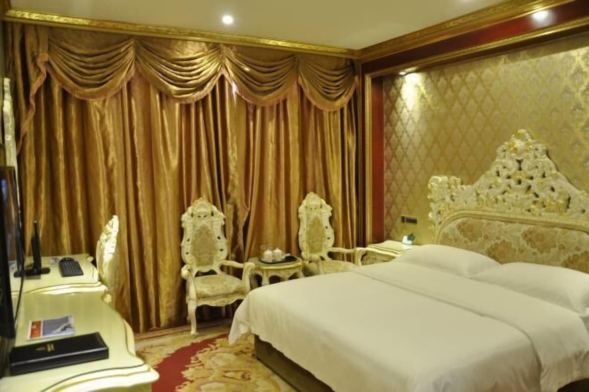A luxurious hotel room with a large bed, two ornate chairs, a desk with a computer, and a red carpet.