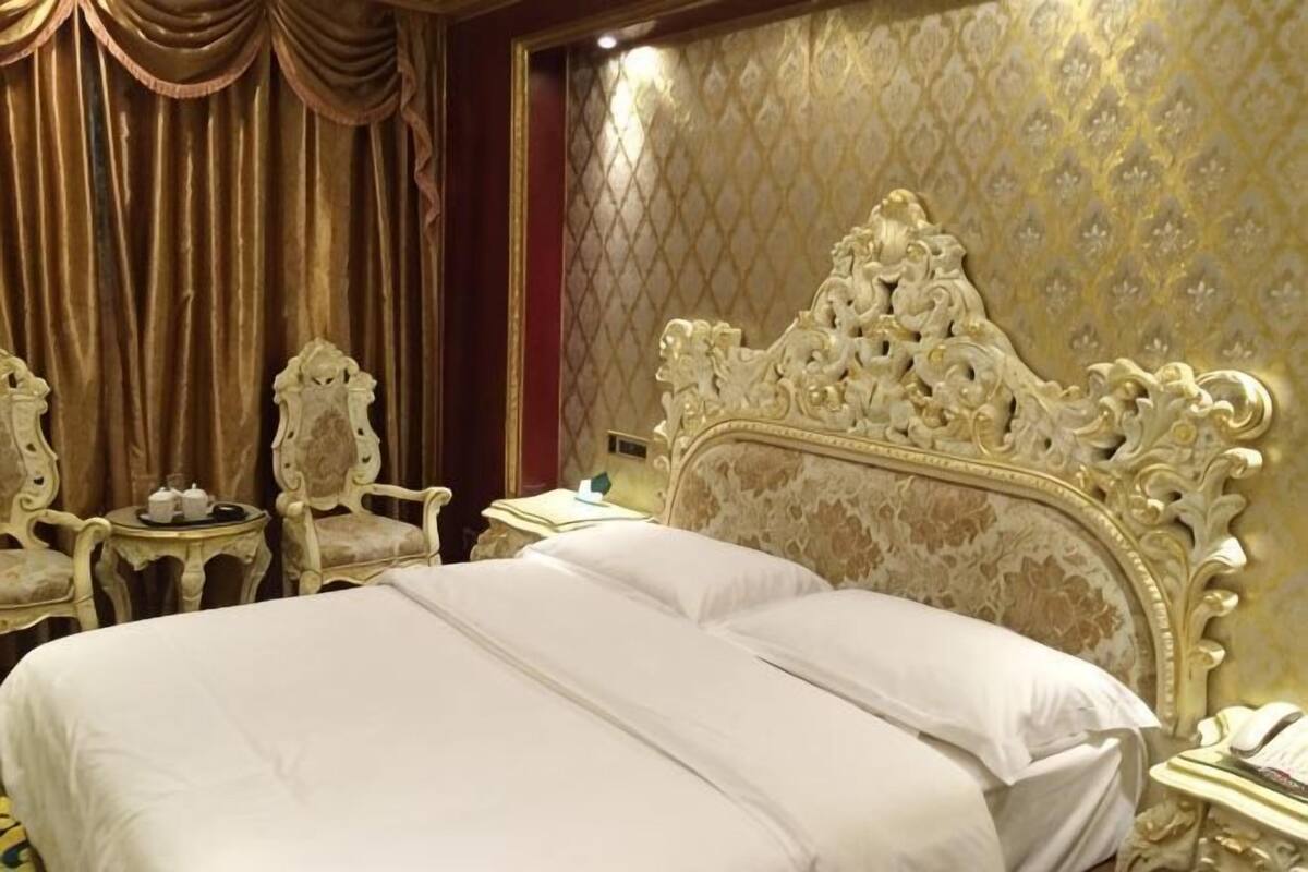 Una cama lujosa con un cabecero decorado, una mesita de noche con lámpara, y una silla con una pequeña mesa.